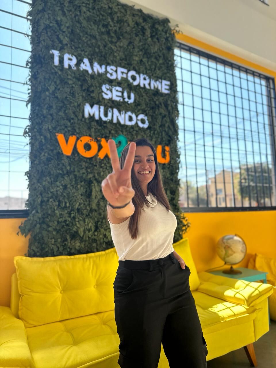 Pietra Rios, fundadora da Social Rios, sorrindo e fazendo sinal de paz em um estúdio com sofá amarelo.